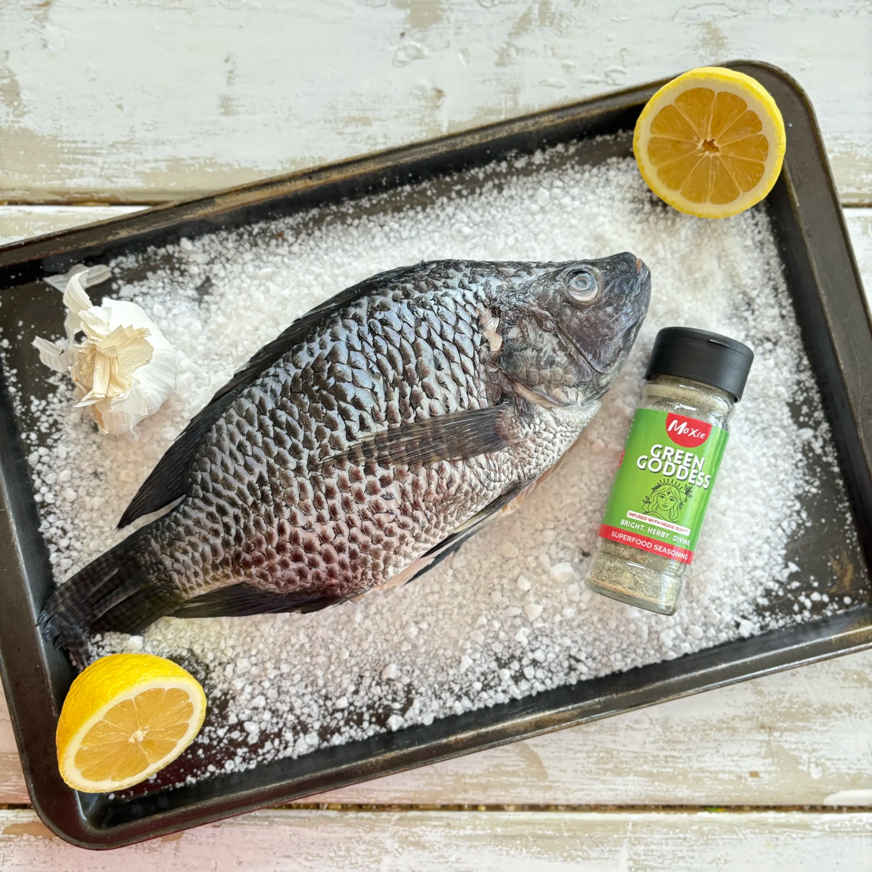 Whole tilapia on a bed of sea salt with Moxie Green Goddess seasoning, garlic, and lemon halves ready for baking