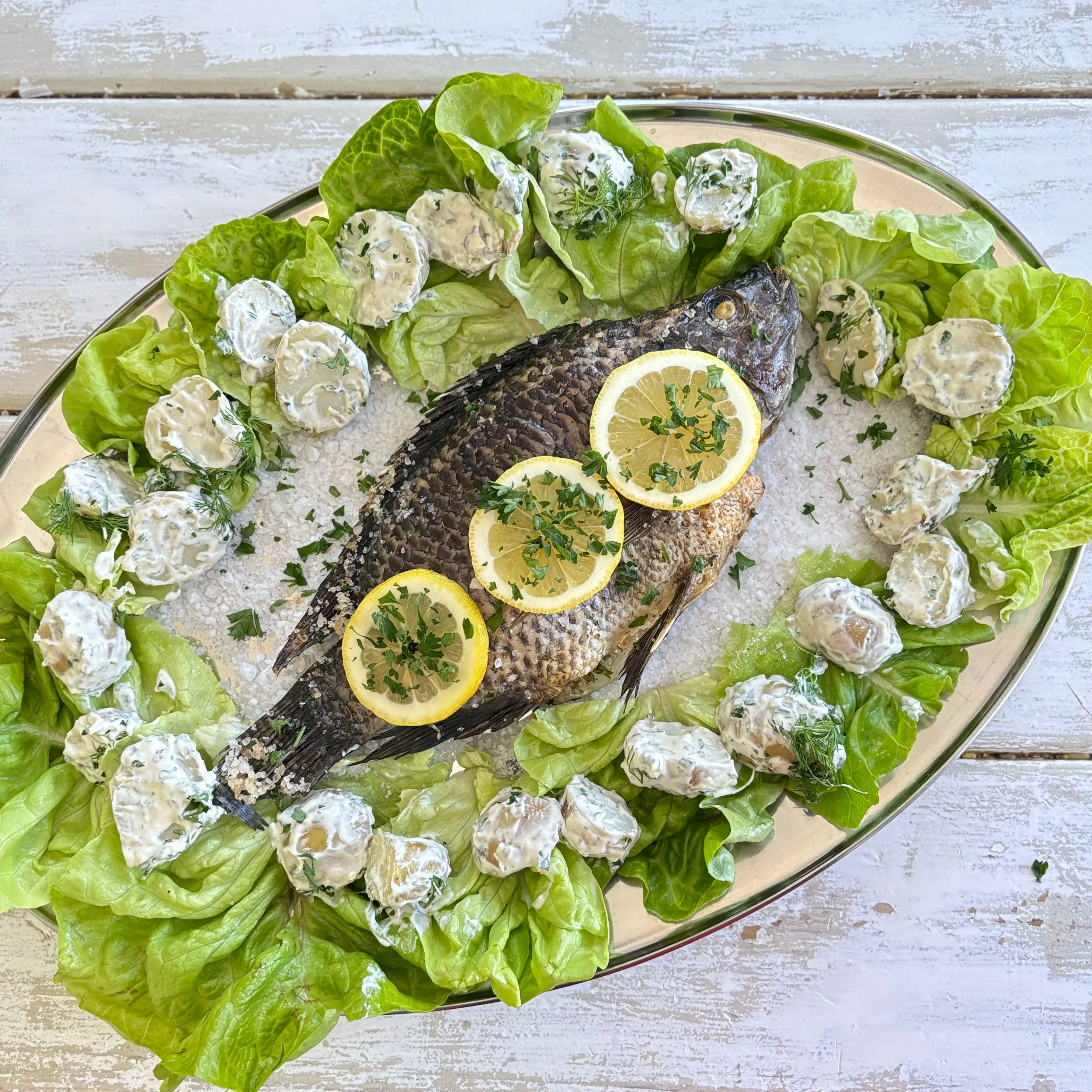 Whole Mediterranean baked tilapia with lemon slices, Green Goddess seasoning, lettuce, and creamy potato salad on a serving platter
